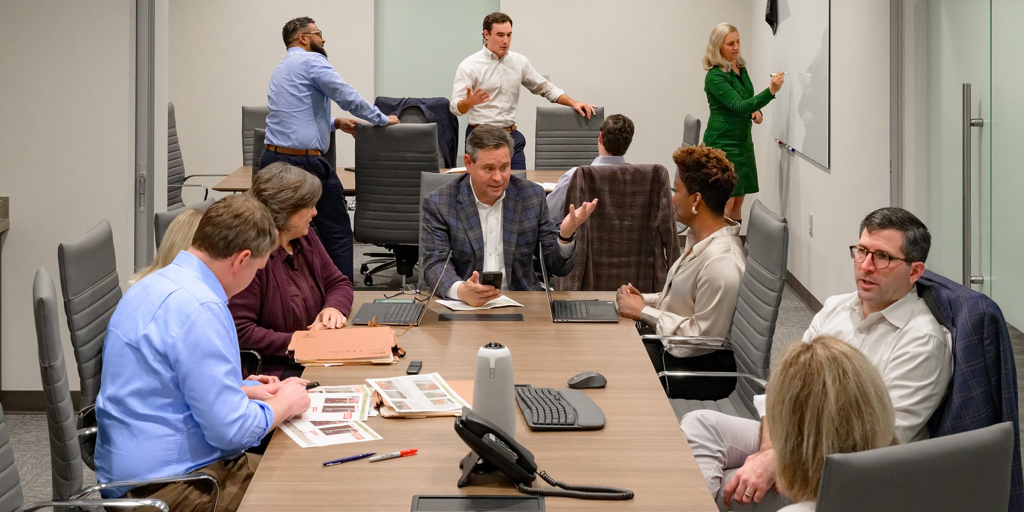 A diverse group of professionals in a modern conference room, engaged in a meeting with laptops, documents, and a whiteboard.